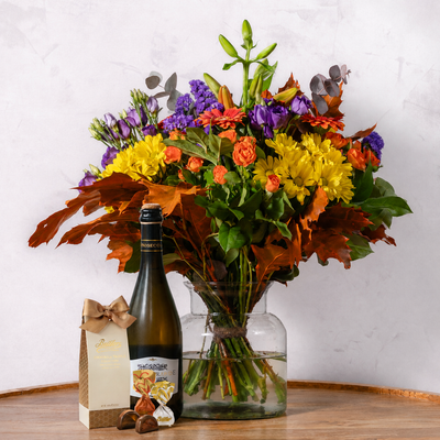 A large autumn floral arrangement in a clear glass vase on a wooden table, styled with a bottle of wine, a gift box, and chocolates against a light textured wall.