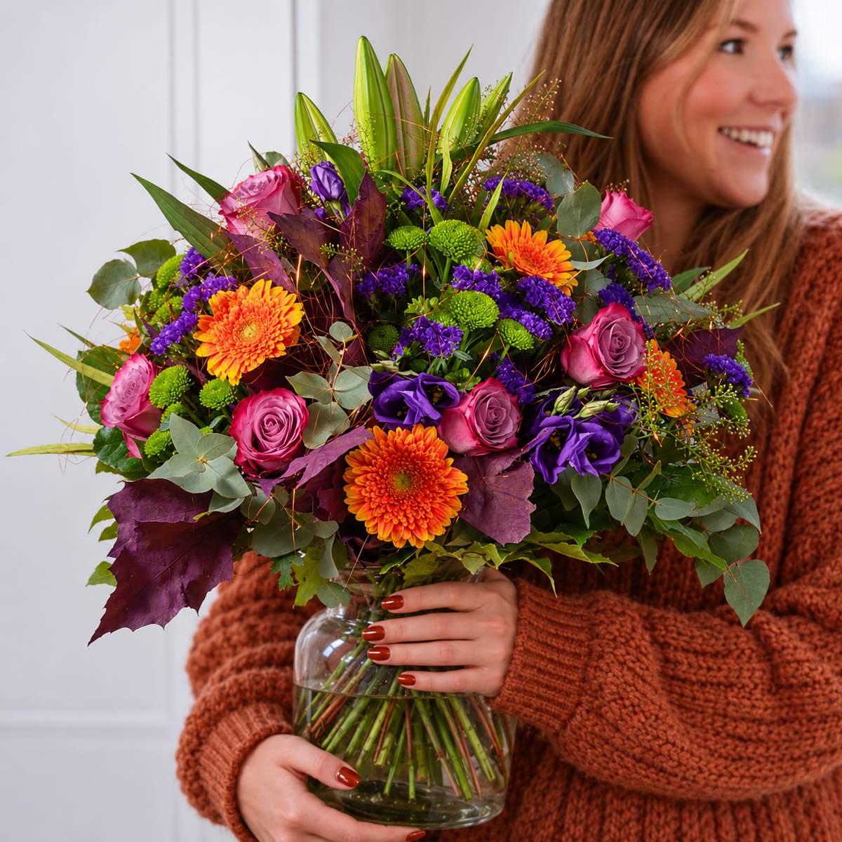 A woman in a rust-brown knit sweater holds a large, vibrant flower bouquet in a clear glass vase. The bouquet features orange gerbera daisies, purple and pink roses, purple lisianthus, green mums, and green foliage.