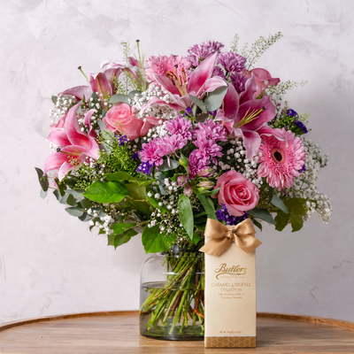 A vibrant floral arrangement of pink lilies, roses, gerbera daisies, and purple chrysanthemums in a clear glass vase with a gold ribbon bow, next to a Butter Channel Triple Collection box on a wooden table against a light gray marble wall.