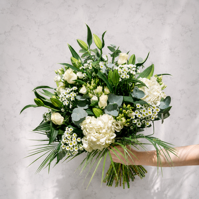 A bouquet of white flowers and green foliage held by a person's arm against a light gray marble-textured background.