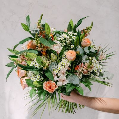 A vibrant floral bouquet held by a person's hand against a light gray marble background. The bouquet features peach roses, white daisies, green foliage, and eucalyptus leaves.