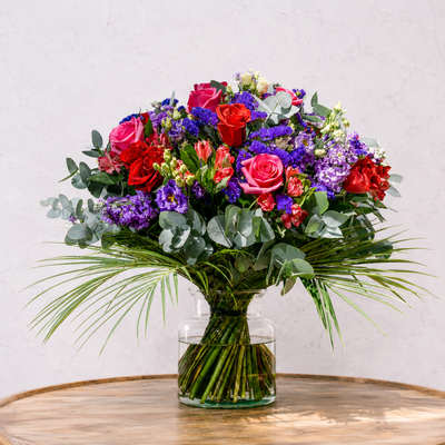 A vibrant artificial flower bouquet in a clear glass vase, featuring red and pink roses, purple and blue filler flowers, green eucalyptus, and palm leaves, displayed on a wooden table against a light textured wall.