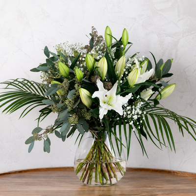Bouquet of white lilies and greenery in a clear vase on a wooden surface with a light gray background