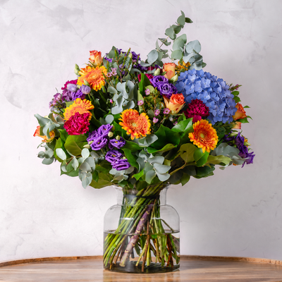 A vibrant, multicolored flower bouquet featuring hydrangeas, gerbera daisies, purple ranunculus, and orange roses, styled with eucalyptus leaves in a clear glass vase on a wooden surface against a light gray marble wall.