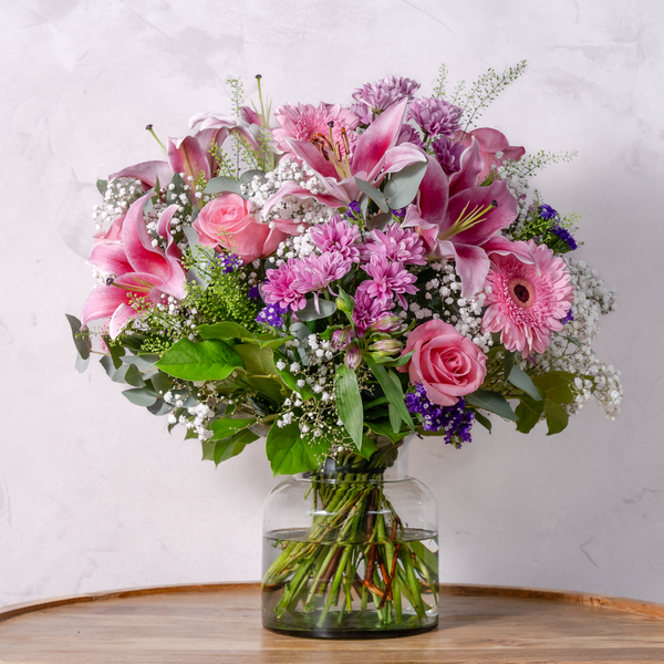 A vibrant hand-tied flower bouquet featuring roses, lilies, gerbera daisies, lisianthus, and other seasonal blooms in a clear vase.