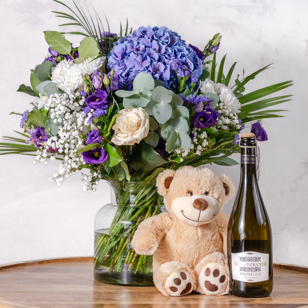 A hand-tied bouquet of fresh flowers featuring blues and whites, accompanied by a plush teddy bear and a bottle of prosecco.