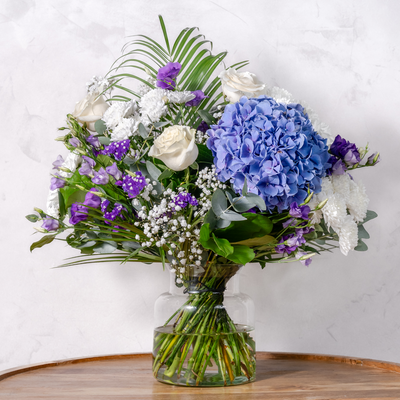 A floral bouquet with a variety of flowers including roses and hydrangeas in shades of blue, purple, and white, arranged in a clear glass vase.