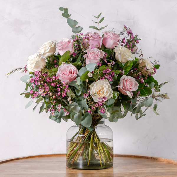 A floral bouquet with a variety of pink and pale cream roses, green leaves, and filler flowers in a clear glass vase, arranged on a wooden surface against a white background.