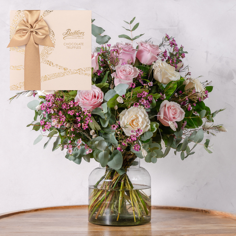 Bouquet of flowers in a glass vase with a box of chocolates on a wooden surface.