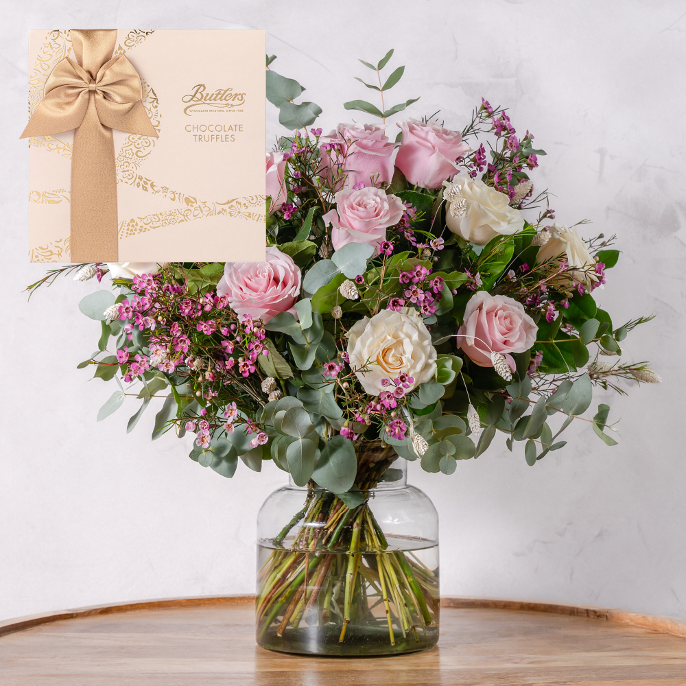 Bouquet of flowers in a glass vase with a box of chocolates on a wooden surface.