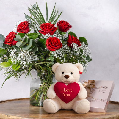 Teddy bear with a heart-shaped 'I Love You' pillow next to a bouquet of red roses on a wooden table.