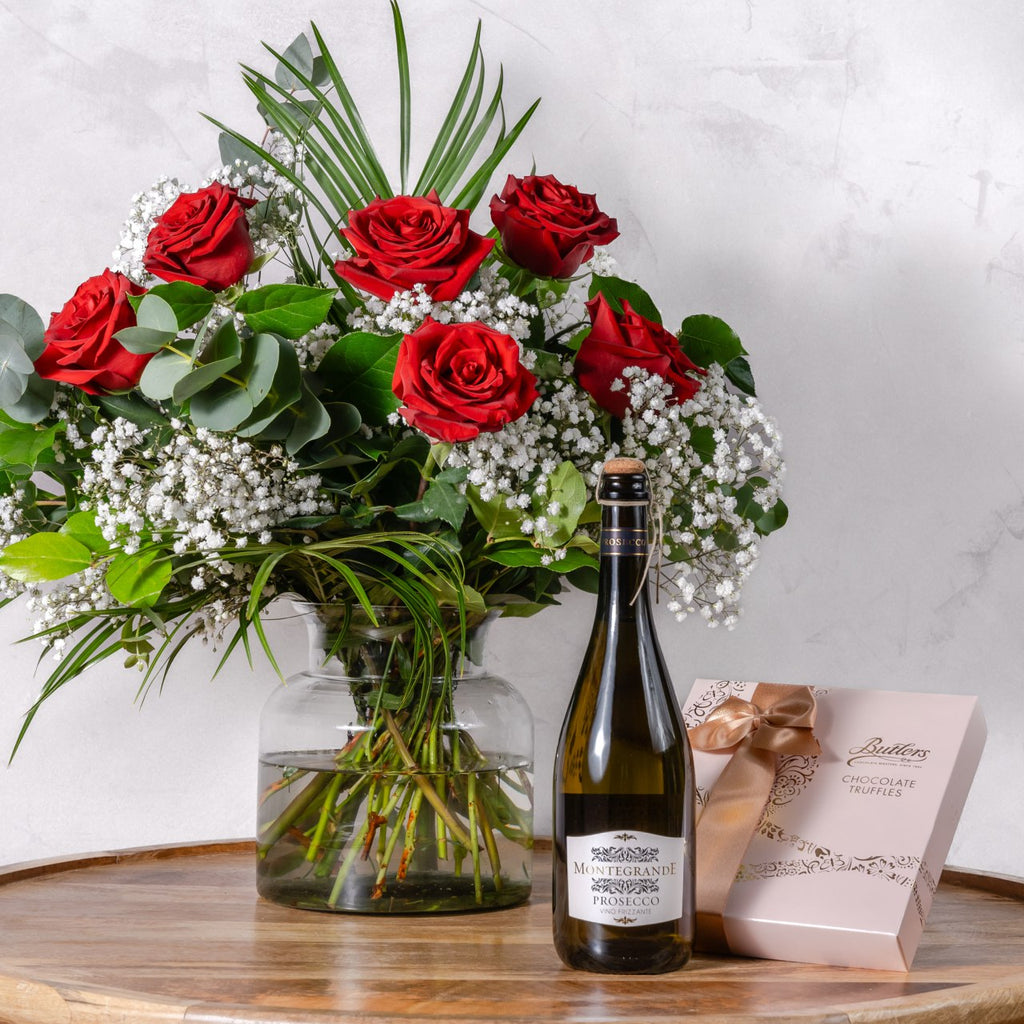 A hand-tied bouquet of red roses, white flowers, and greenery in a clear vase accompanied by a bottle of Prosecco and a box of chocolates.