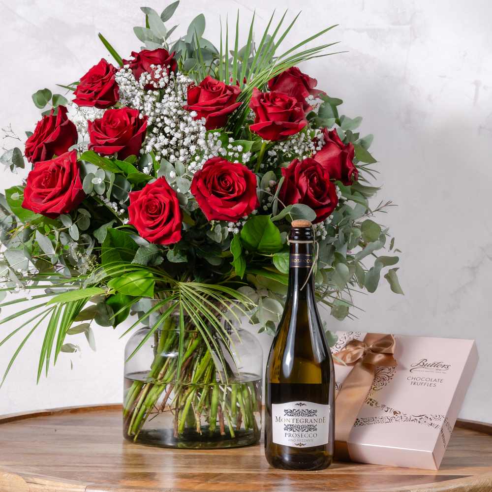 A dozen red roses in a clear vase, accompanied by a bottle of prosecco and a box of chocolates.