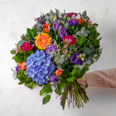 A vibrant mixed floral bouquet held by a hand wearing a beige sleeve, featuring a large blue hydrangea, purple lisianthus, orange gerbera daisy, and pink carnations with green foliage, set against a light gray marble background.