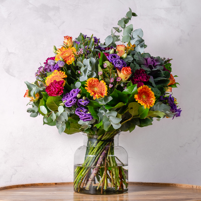 A colorful bouquet of flowers including red roses, orange daisies, and purple flowers with green foliage, arranged in a clear vase.