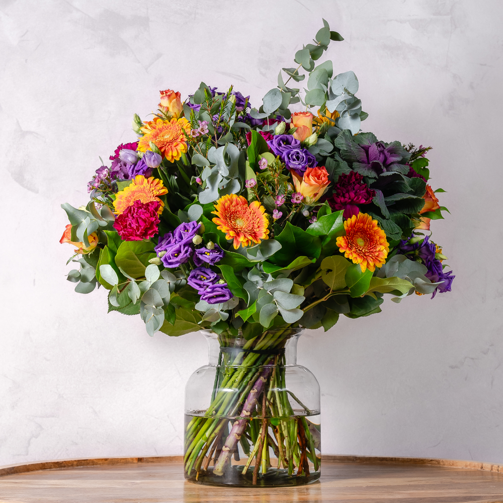 A colorful bouquet of flowers including red roses, orange daisies, and purple flowers with green foliage, arranged in a clear vase.