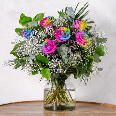A bouquet of rainbow-colored roses with green leaves and white filler flowers in a clear glass vase.