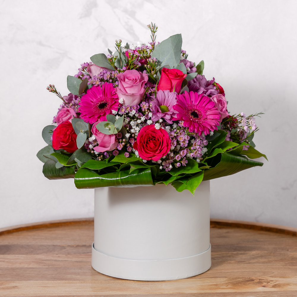 A colorful floral arrangement of flowers such as roses and gerberas, presented in a white cylindrical hat box.