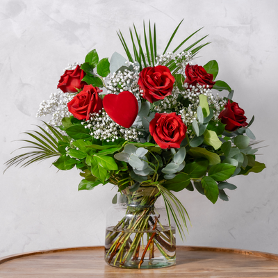 Bouquet of red roses with a heart-shaped object in a clear vase on a wooden surface.