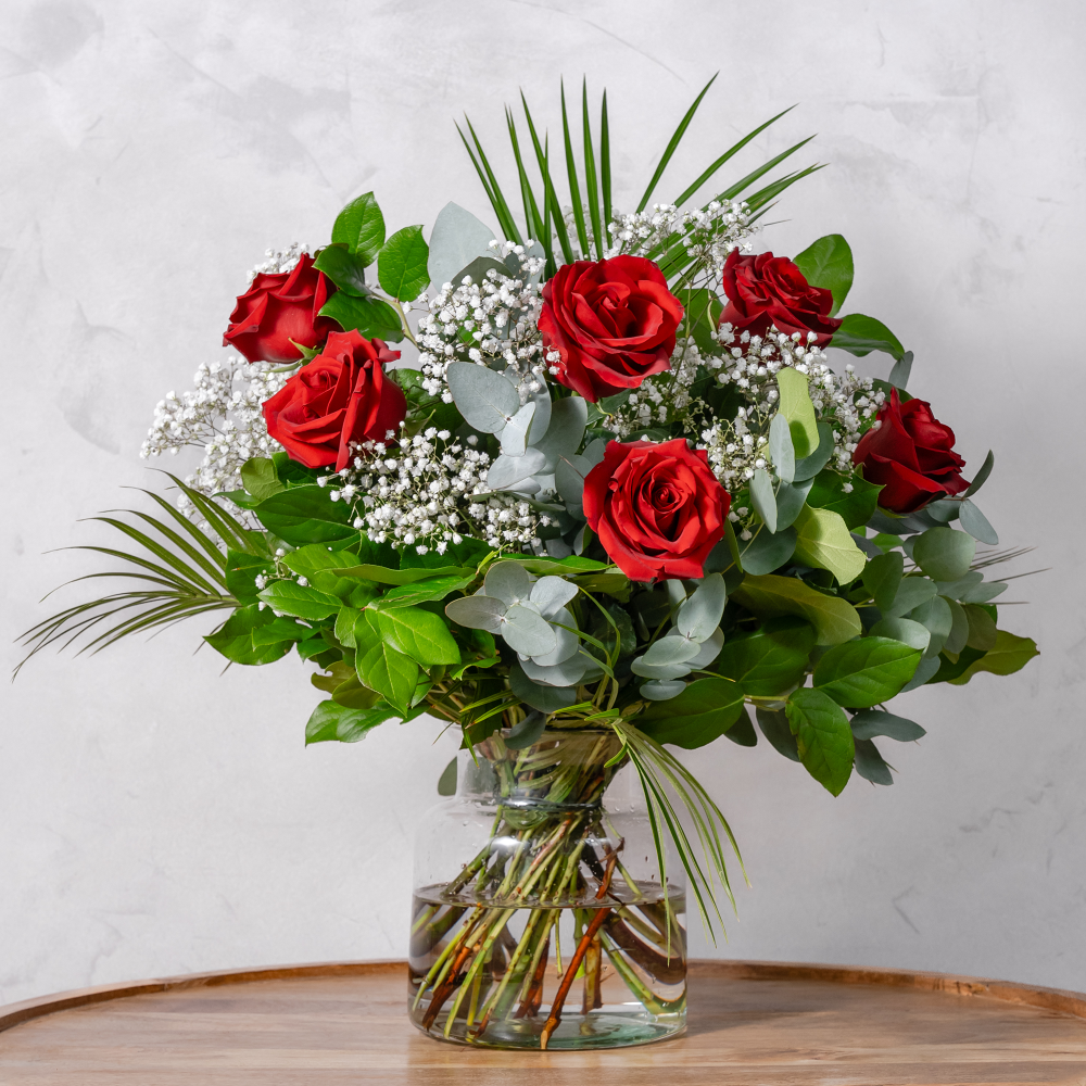 A vase of half dozen red roses with green leaves and white floral accents.