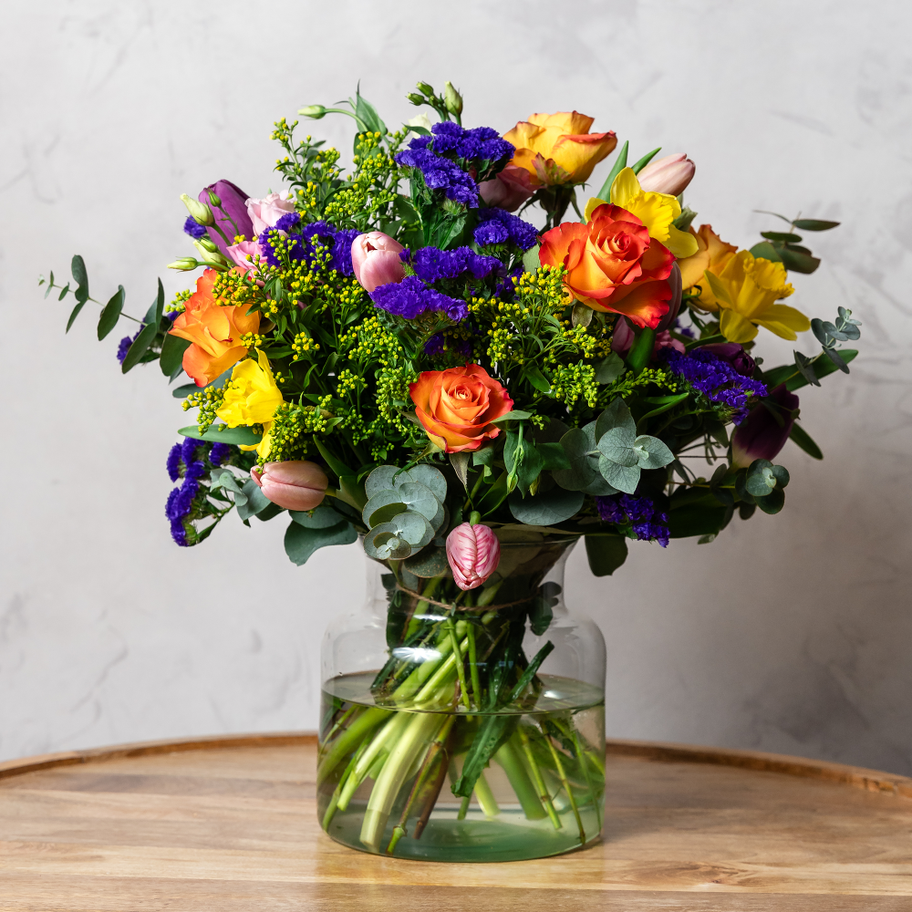 A vibrant bouquet of handpicked blooms including roses, gerbera daisies, and lisianthus in a clear vase, arranged in radiant seasonal colors.