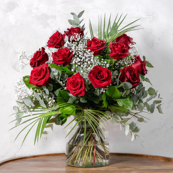 A dozen red roses arranged in a clear glass vase with green leaves and white floral accents.