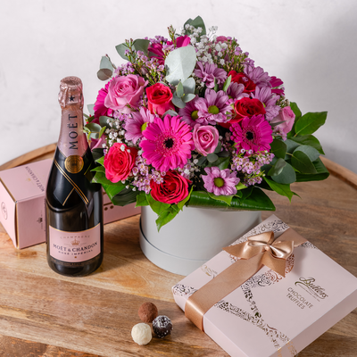 A gift bundle arrangement featuring a bouquet of pink and red flowers in a hatbox, accompanied by a bottle of Moet & Chandon Rosé champagne and a box of chocolates.